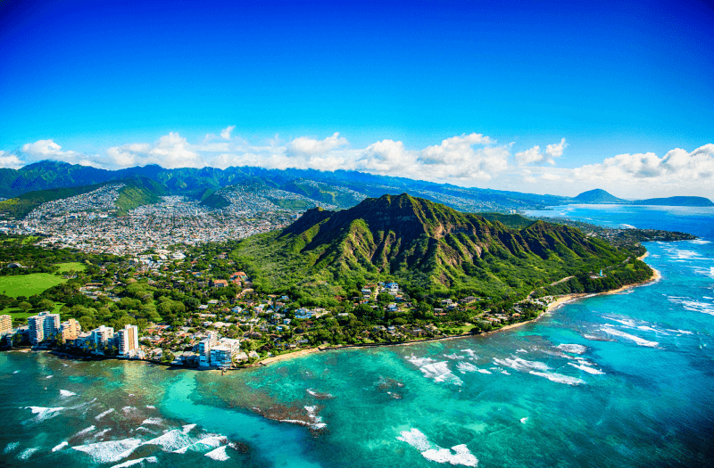 Aerial view of the coast surrounded by buildings, vegetation and a mountain, with mountains and clear sky in the background