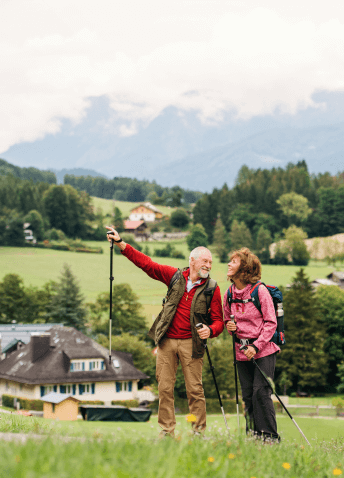 Two people on the meadow with hiking poles in their hands, with buildings, trees and clear sky in the background