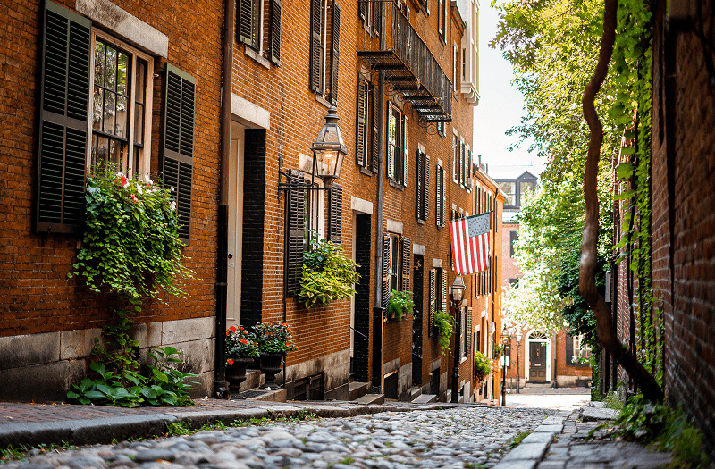 A narrow cobblestone street in Boston with lush flower boxes lining the historic buildings windows.