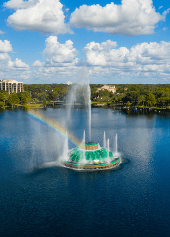 A vibrant rainbow arches over a fountain in an Orlando area lake, with hotel buildings and lush trees in the background.