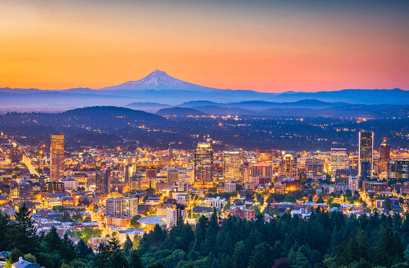 Portland's illuminated skyline at sunset with Mount Hood against the orange and purple sky in the distance.