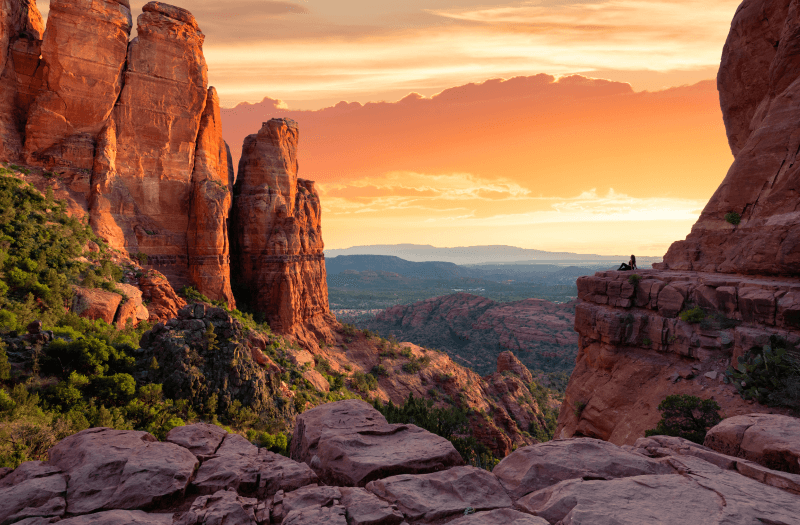 Rocky red canyon landscape at sunset with dramatic clouds and a person sitting on a ledge, overlooking the distant valley and mountains.