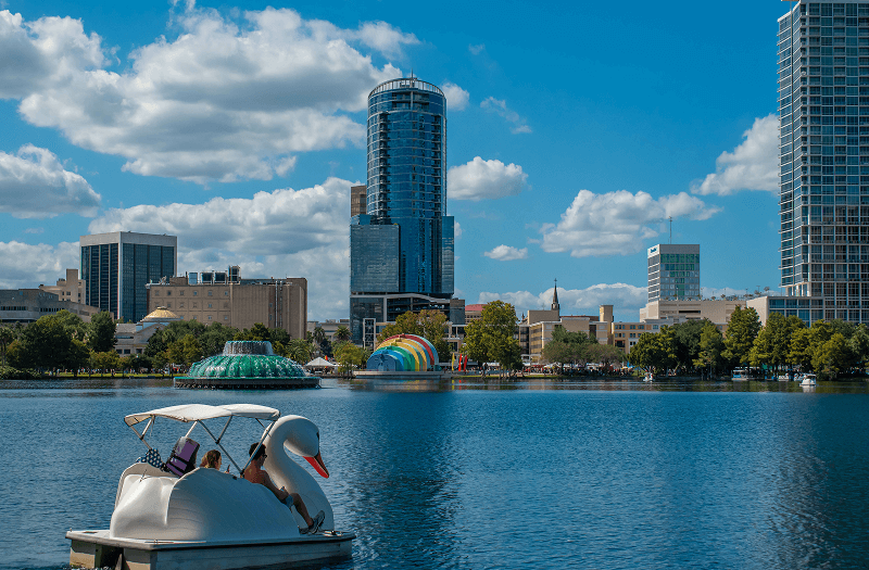 A couple riding a swan-shaped paddle boat in a lake with Orlando downtown in the background.