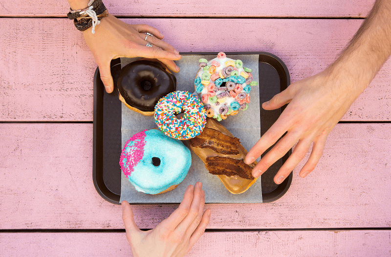 A tray of colorful assorted donuts on a pink table with three hands reaching for them.