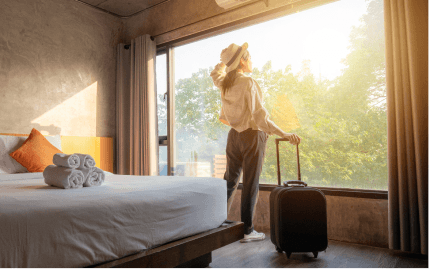 A woman with a black rolling suitcase standing next to a large hotel window and looking out.