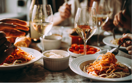 A close-up of a table where people are eating with several glasses of white wine and plates of pasta.