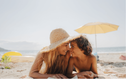 A smiling mom and son looking nose to nose while lying on a sandy beach on a clear day.