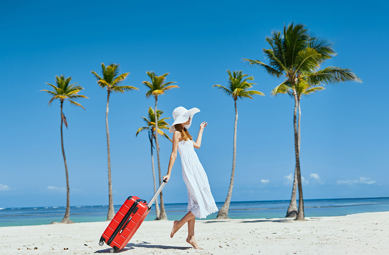 A woman with a white dress and white hat toting a red suitcase along a beautiful sandy beach on a clear day.
