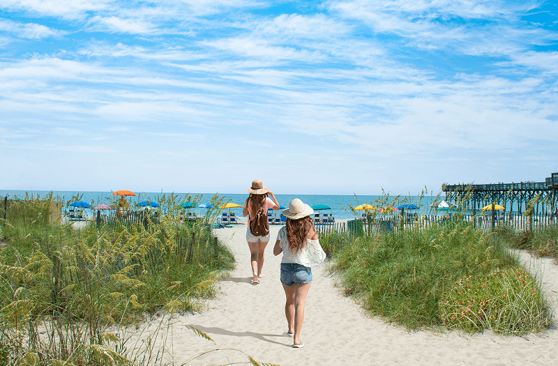 Two ladies with hats walking down a small path toward the sandy shores of Myrtle Beach.