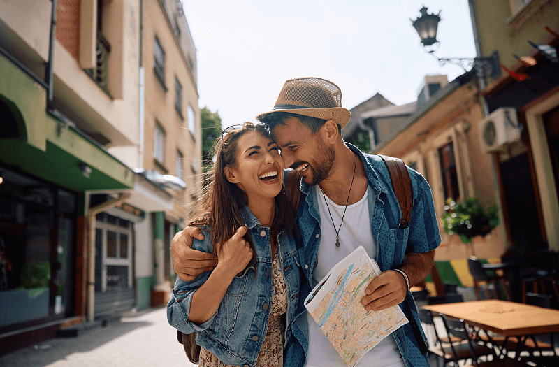 A smiling man with his arm around a smiling lady as they walk along a city street holding a map.
