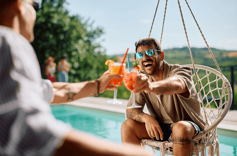 A smiling man toasting a cocktail with his friend while sitting poolside on a summer day.