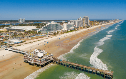 Aerial view of the Daytona Beach Boardwalk and Pier in the mid-morning with people scattered along the calm shores.