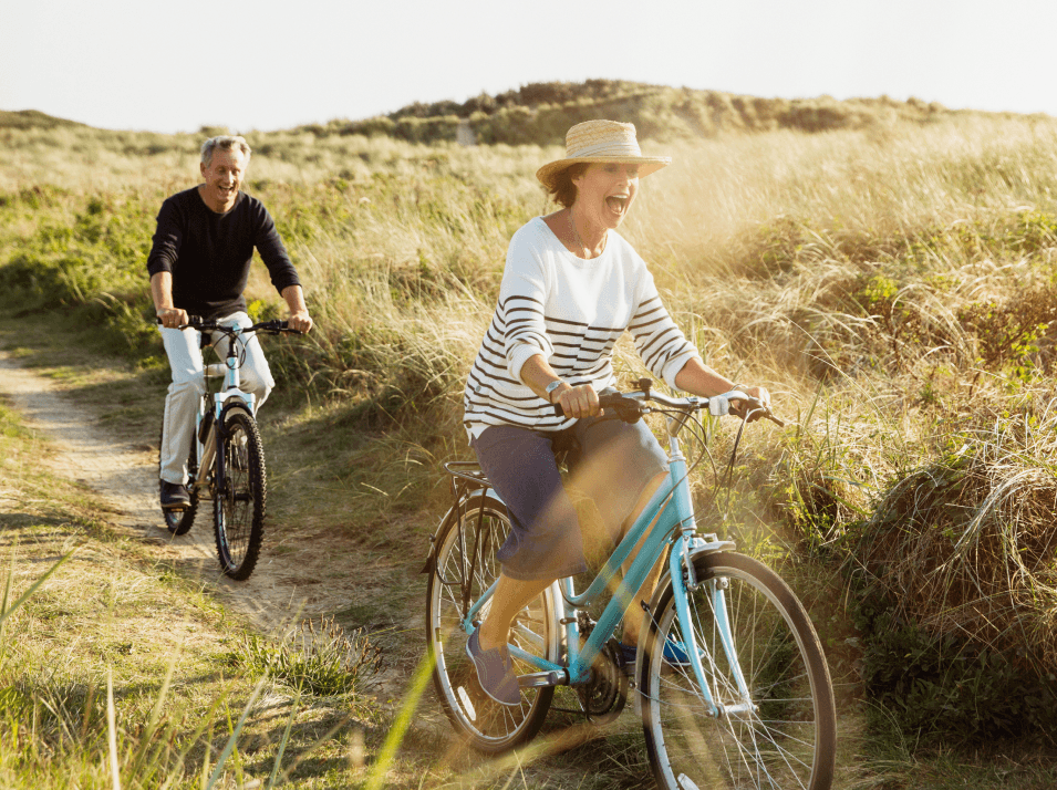 A happy man and woman riding bikes along a dirt path, surrounded by green hills and vegetation.