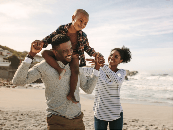 A son sitting on his dad’s shoulders as they walk along the beach next to mom on a chilly day.