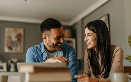 A smiling man and woman looking at one another while they sit at a table with a computer.