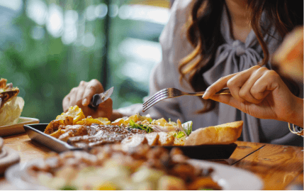 A close-up of a woman’s hands holding a knife and fork above a delicious-looking plate of food on a wooden table.