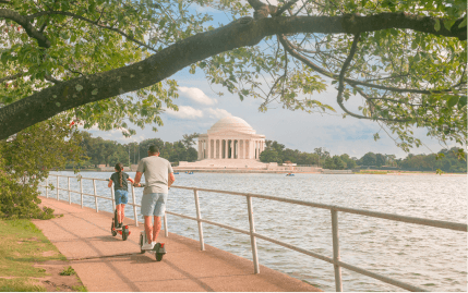 A dad and daughter riding scooters along a path next to the Tidal Basin, across from the Jefferson Memorial.