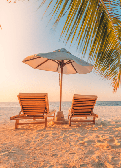 Two wooden lounge chairs and a white umbrella sitting on a white sand beach under a palm tree during sunset. 