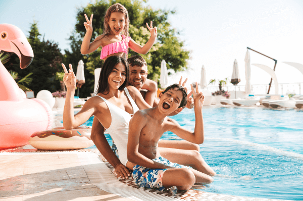 Two adults and two children sitting on the edge of a pool smiling and waving