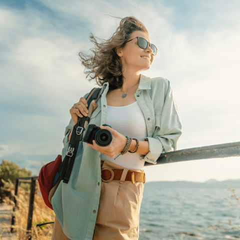 Person leaning on a railing, holding a camera with the sea and vegetation in the background