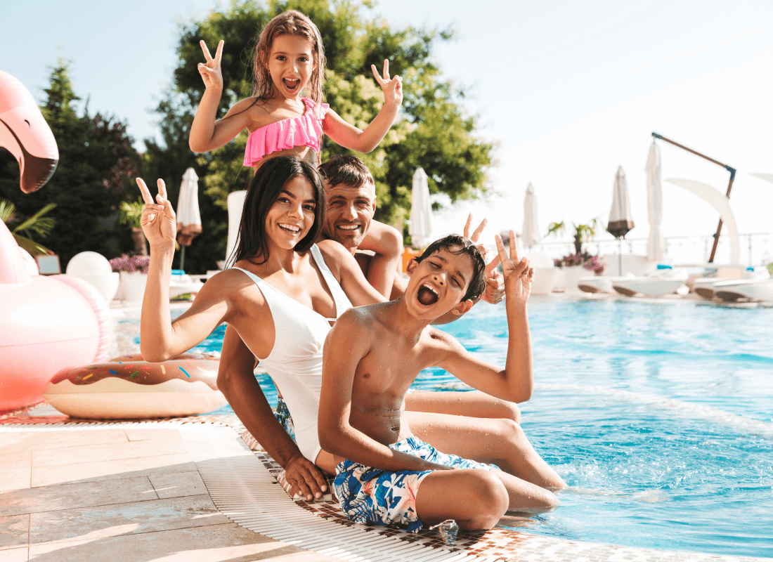 Two adults and two children sitting on the edge of a pool smiling and waving