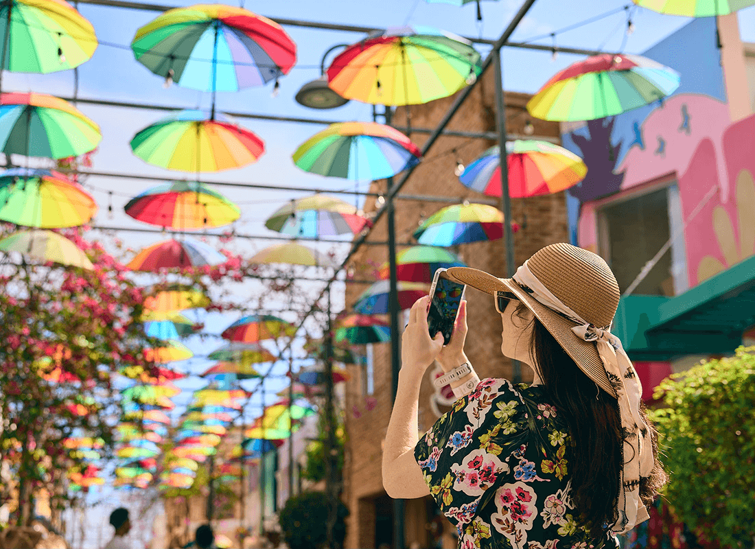 A lady in a sun hat taking a photo with her phone of her surroundings: brightly colored umbrellas hanging over the street