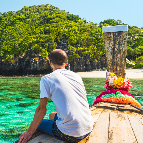 Rear view of a person sitting in a wooden boat, with a beach and wooded mountain in the background