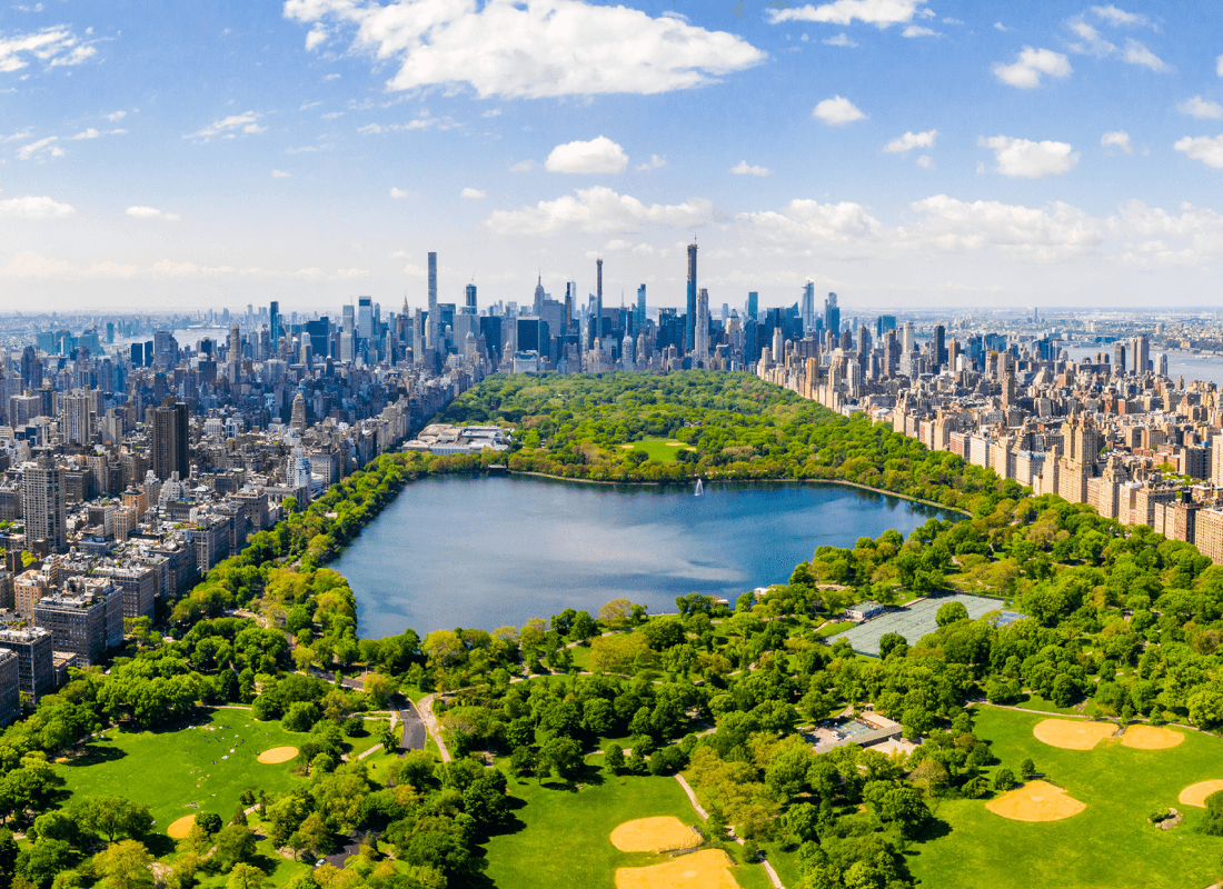 Aerial view of Central Park in New York City with skyscrapers in the background, a large reservoir, green spaces, and pathways visible under a partly cloudy sky.