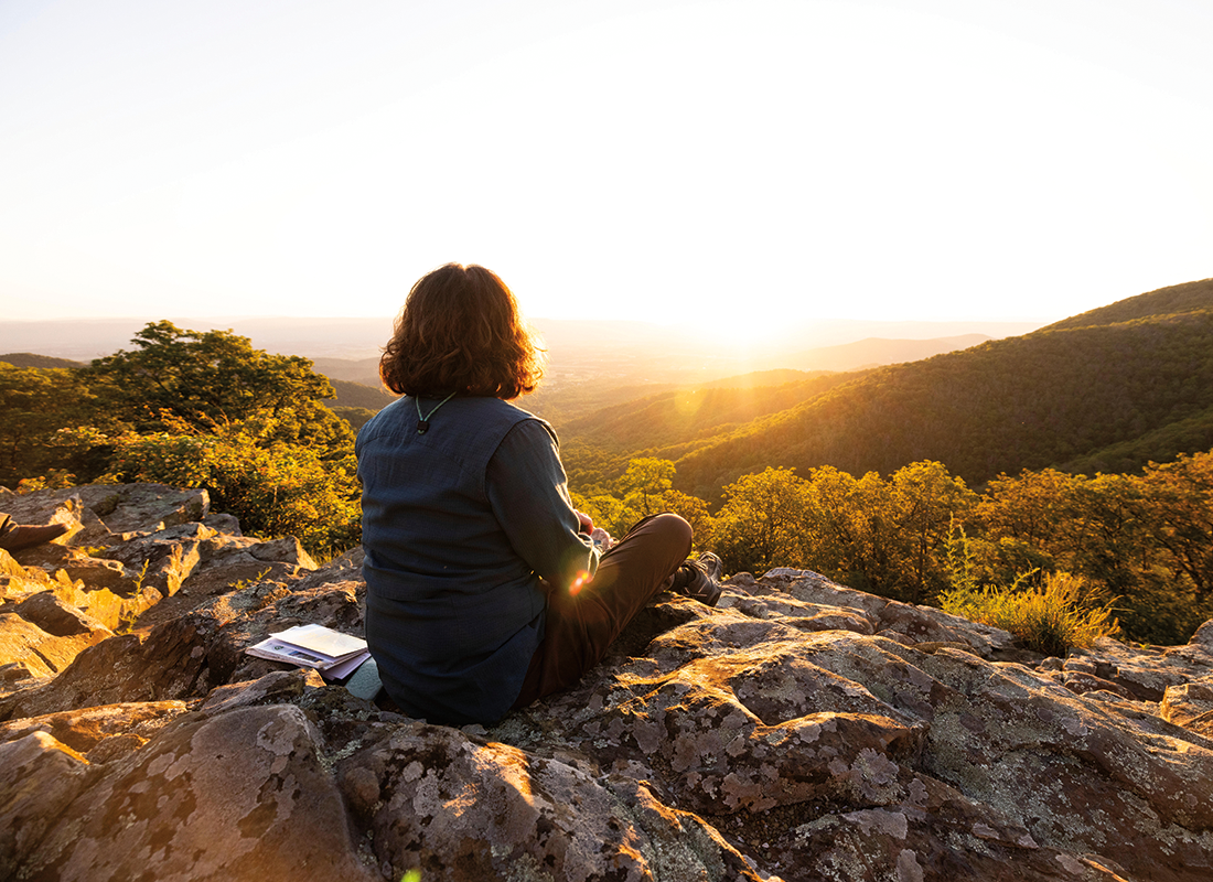 View from behind of a person enjoying the sunset while sitting on a rock, with forested mountains in the background