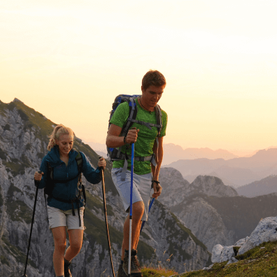 A man and woman hiking atop a mountain with the sun setting in the distance.