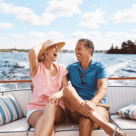 A couple enjoying a sunny day on a boat while the woman holds on to her hat.