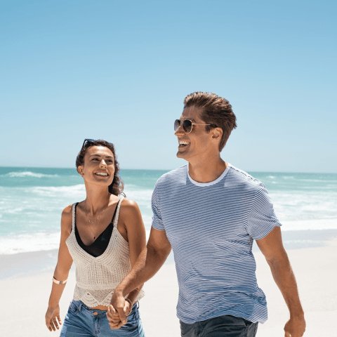 A man with a blue striped shirt and sunglasses holding hands with a lady in a tank top while they walk along the beach on a bright afternoon.