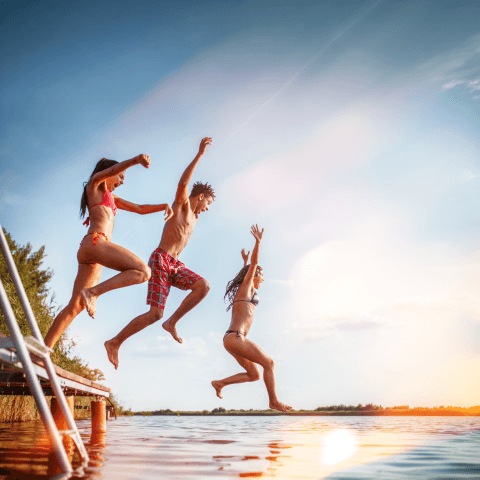 Three friends in swimwear jumping off a wooden dock into a calm lake under a bright, sunny blue sky.