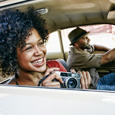 A smiling woman riding a car while taking photos with the windows down.