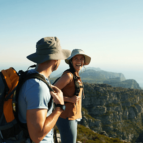 A man and woman wearing backpacks and hats, looking at one another while they stand atop a mountain on a clear day.