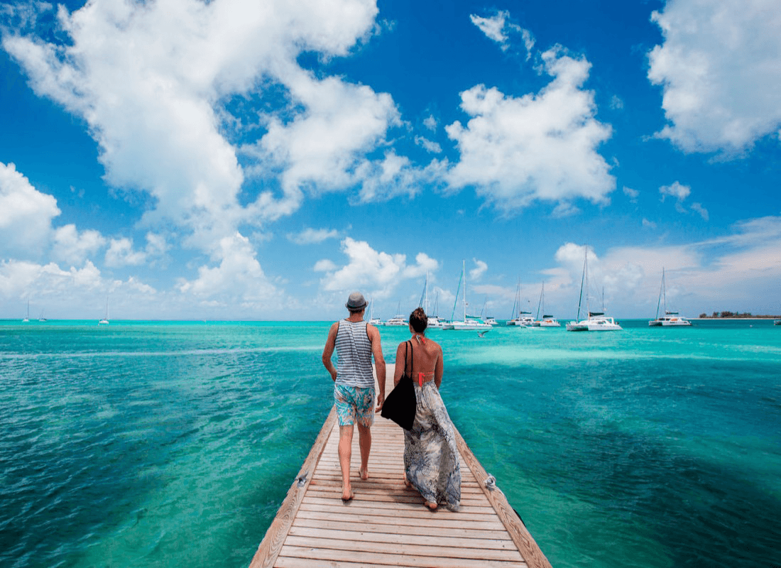A couple walking on a dock towards blue green waters with boats docked in the distance.