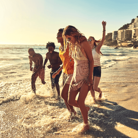 A group of cheerful young friends splashing through ocean waves along a sunny beach shoreline.