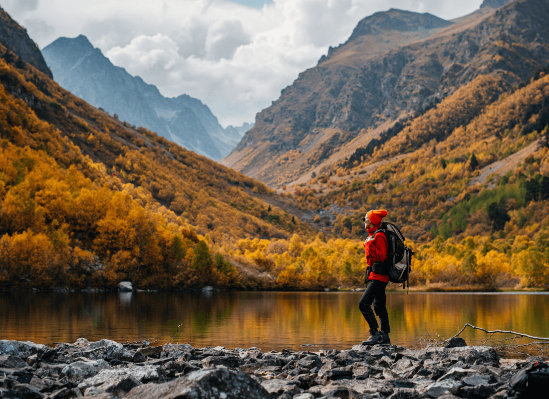 A hiker wearing a red jacket and orange beanie standing by a lake surrounded by autumn trees and rugged mountains.