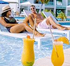 A couple lounging on beach chairs at a resort pool with tropical drinks in the foreground
