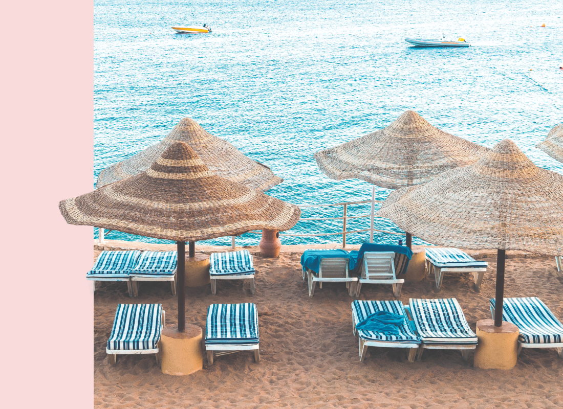 Blue striped lounge chairs with wispy umbrellas next to beautiful waters with a few boats sailing in the distance.