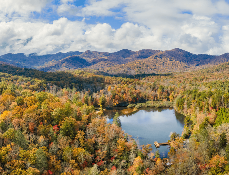 A scenic autumn overlook of a small lake with a wooden dock nestled in a valley near Asheville, North Carolina, surrounded by vibrant fall foliage and rolling blue mountains.