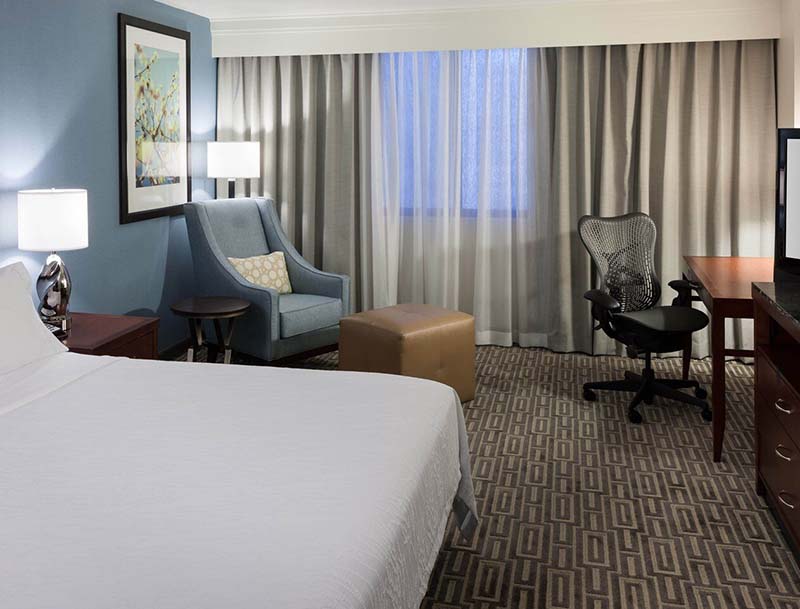A cozy well-lit hotel room featuring a king-size bed, a desk with an office chair, and a sitting area by the window.