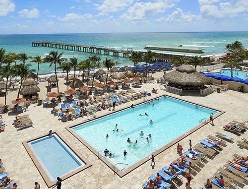 A bird's-eye view of resort guests enjoying the pool and deck area, with a palm-dotted sandy beach and azure ocean waters visible in the distance.
