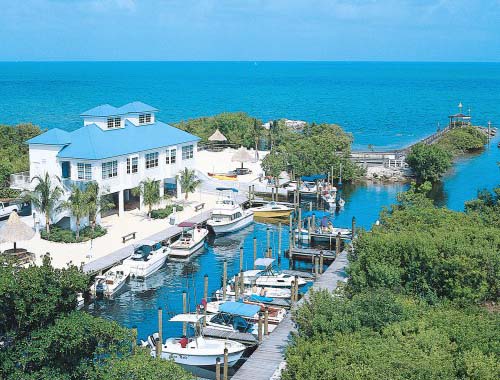 Aerial view of Ocean Pointe Suites in Key Largo, featuring a blue-roofed building and a marina filled with boats surrounded by mangroves and turquoise water.