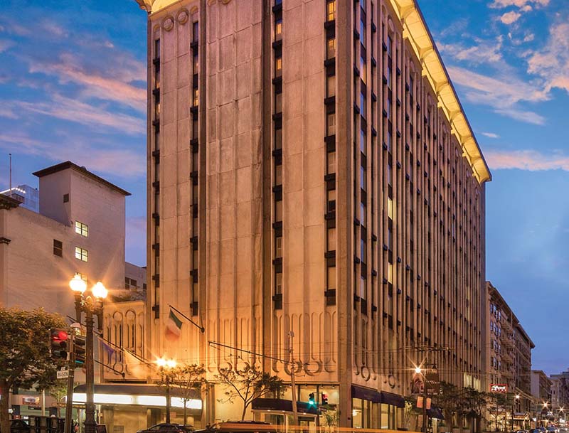 A street corner view of the illuminated hotel rising into the dusky sky.