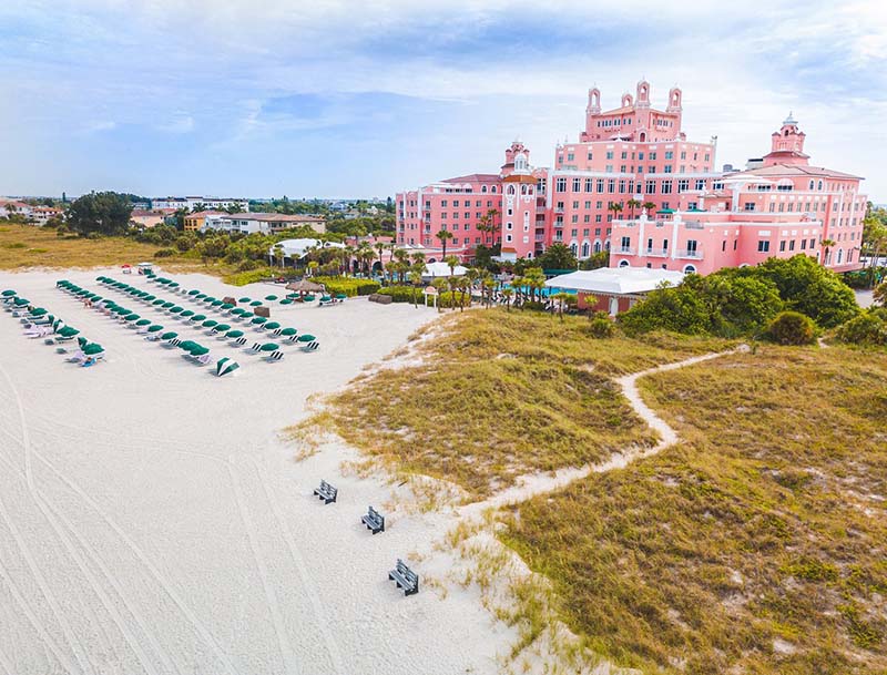 An aerial view of the resort's signature pink facade and the sandy shore lined up with lounge chairs and umbrellas. 