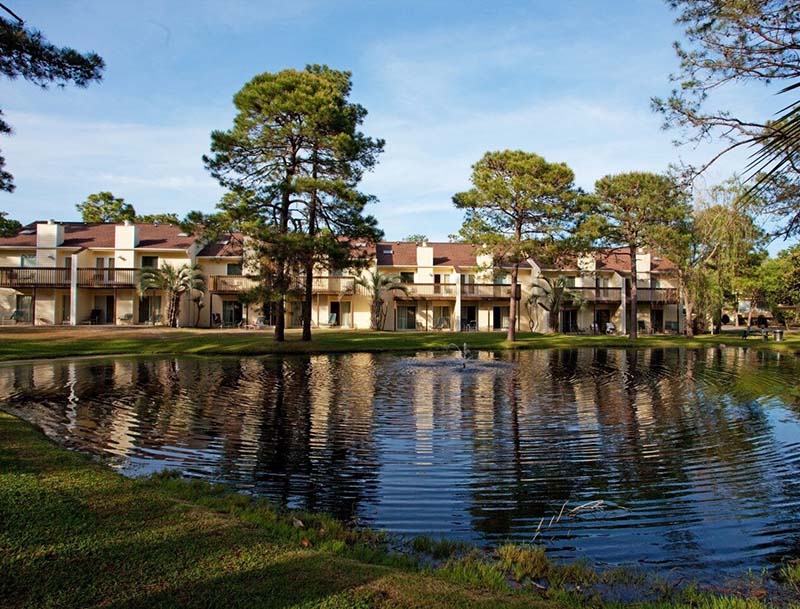 Two-story resort buildings surrounded by lush trees with a serene lake in the foreground.
