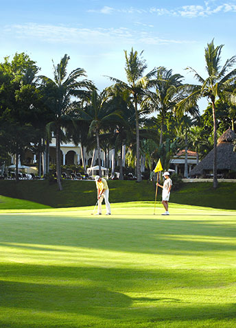 Two people are on a golf green; one is putting while the other holds the flagstick. 