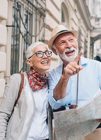 An older couple stands outdoors, smiling and holding a map. Both appear to be enjoying sightseeing.
