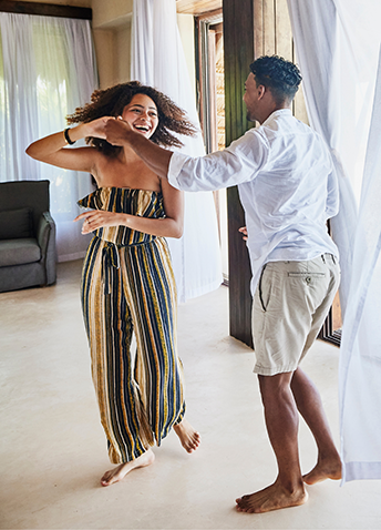 A smiling couple dances barefoot together inside a brightly lit resort room framed by sheer white curtains.